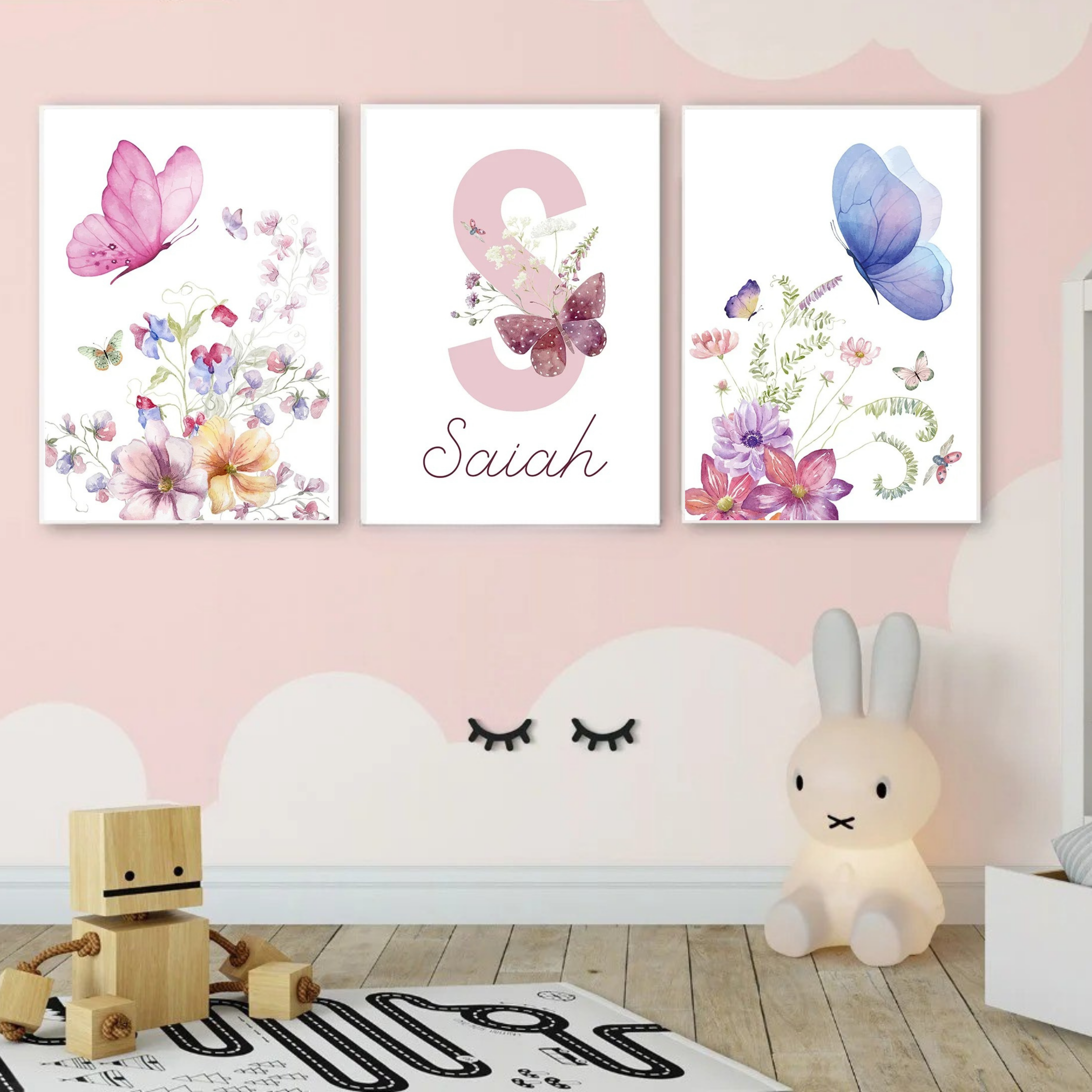 Triptyque d’affiches murales florales avec papillons pastel, installé dans une chambre bébé aux murs roses, avec jouets en bois, tapis graphique et décoration douce et poétique.
