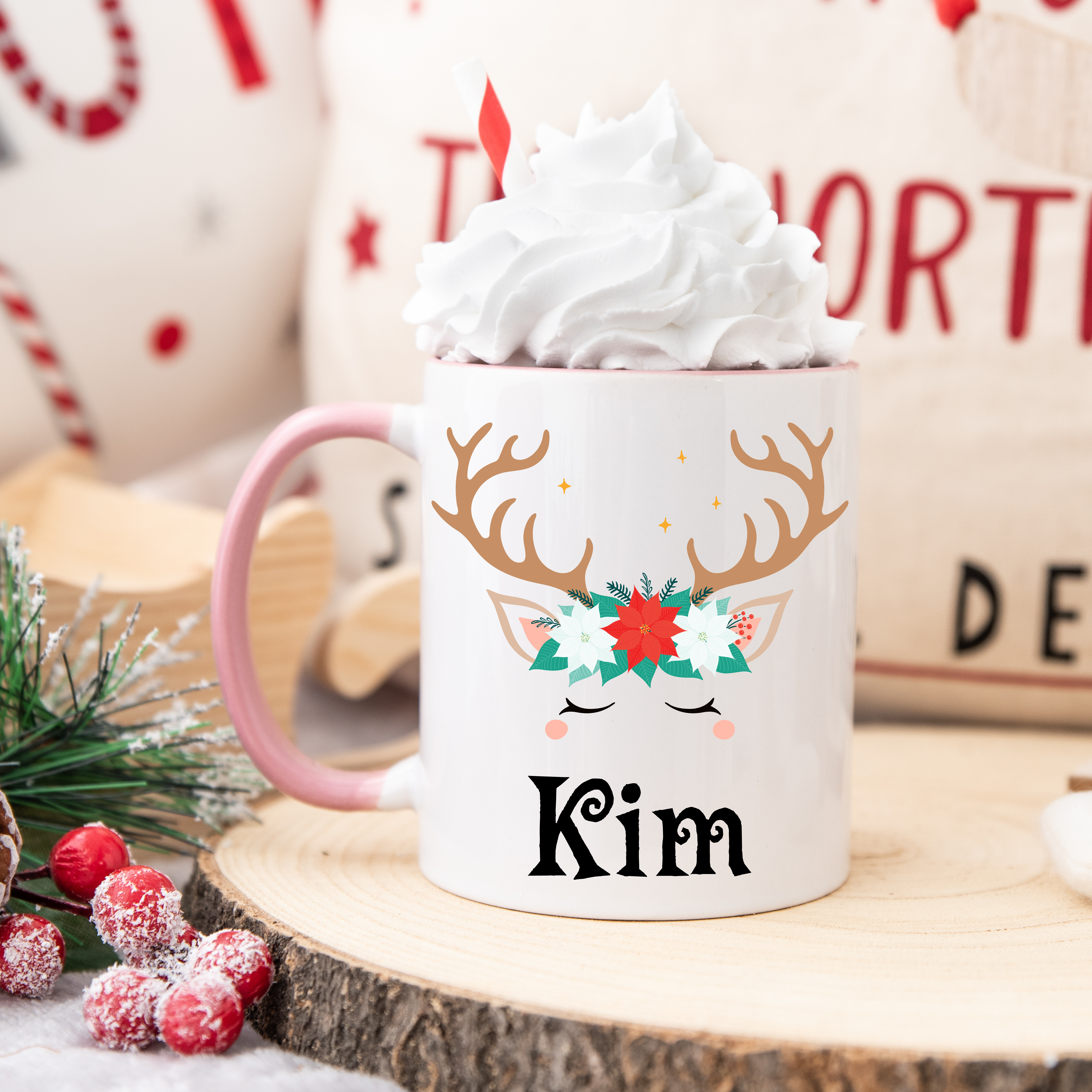 Mug de Noël avec motif renne et couronne florale, anse rose et intérieur rose, posé sur une table avec chantilly et décoration festive.