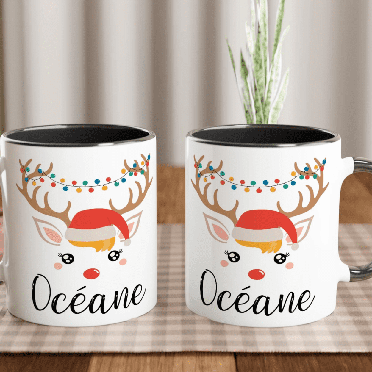 Mug de Noël avec visage de renne, bonnet rouge et guirlande lumineuse sur les bois, anse noire et intérieur noir.