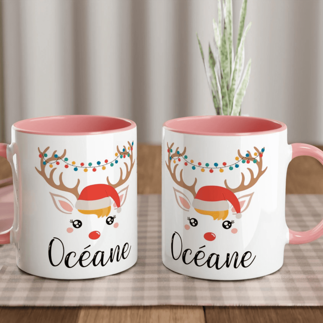 Mug de Noël illustré d’un renne festif avec bonnet rouge et guirlande lumineuse, anse rose et intérieur rose.