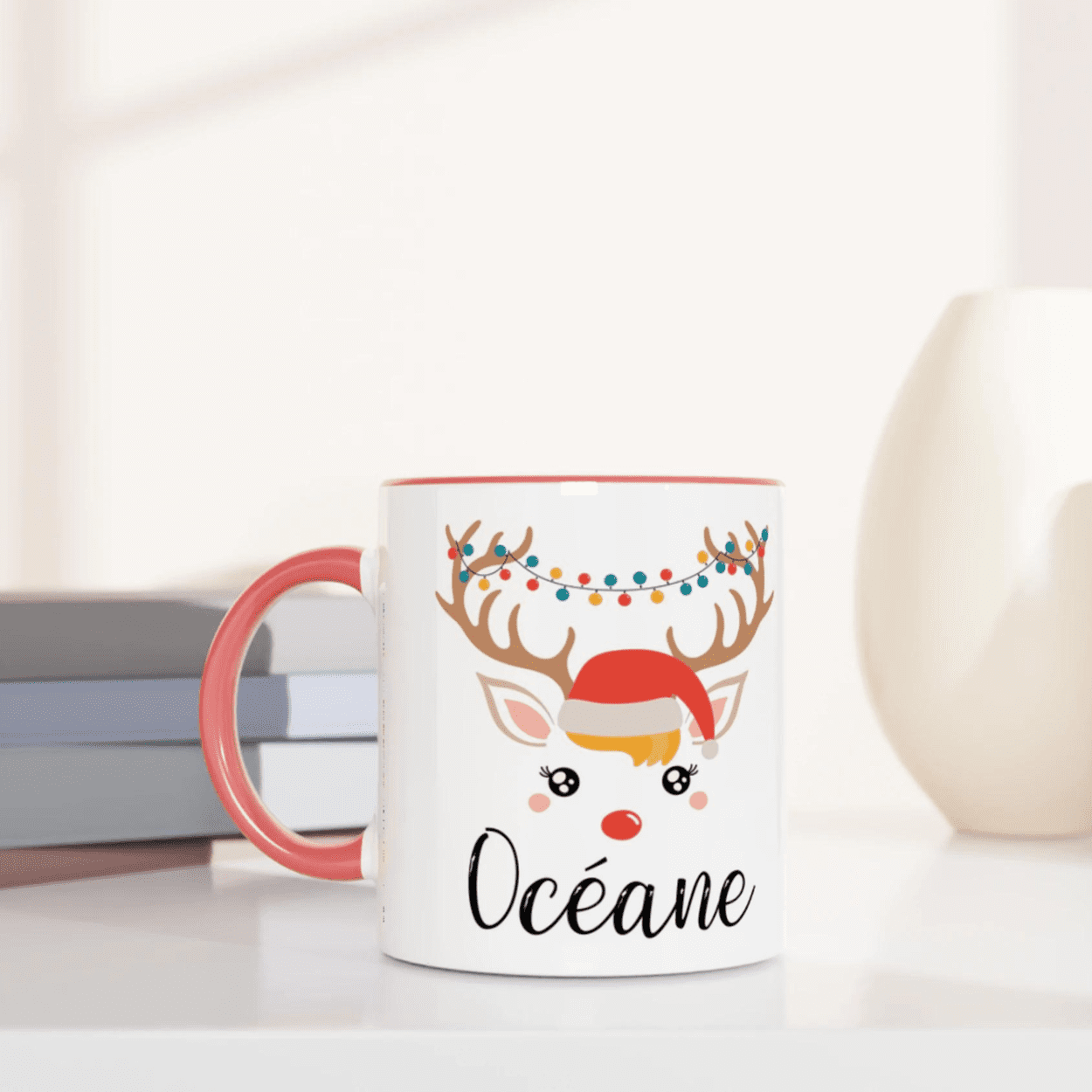 Mug de Noël illustré d’un renne festif avec bonnet rouge et guirlande lumineuse, anse rose et intérieur rose.