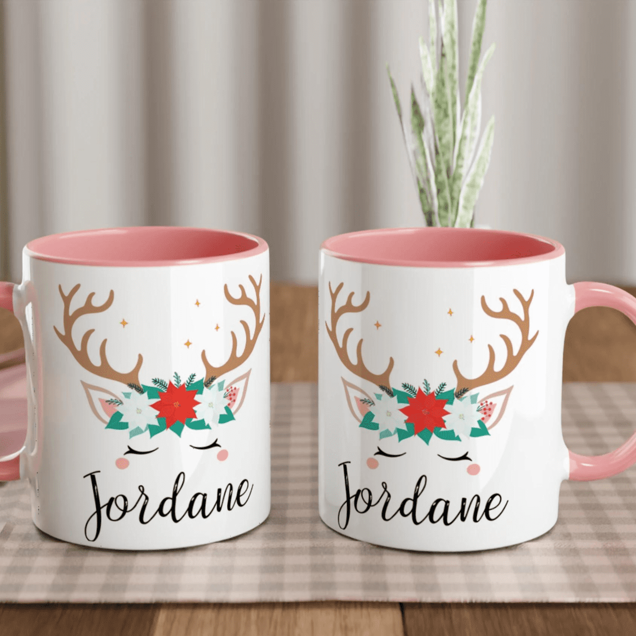 Mug de Noël avec motif renne et couronne florale, anse rose et intérieur rose, posé sur une table avec chantilly et décoration festive.