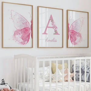 Triptyque d’affiches murales avec ailes roses à l’aquarelle et lettre décorative centrale ornée de fleurs, installé au-dessus d’un lit bébé blanc dans une chambre douce et élégante.