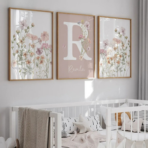 Triptyque d’affiches murales florales à l’aquarelle avec une lettre décorative centrale, accroché au-dessus d’un lit bébé blanc dans une chambre douce et lumineuse aux tons naturels.