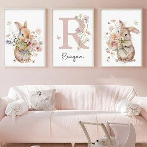 Triptyque d’affiches murales avec lapins illustrés à l’aquarelle et lettre décorative entourée de fleurs, installé au-dessus d’un canapé clair dans une chambre d’enfant à l’ambiance douce et élégante.